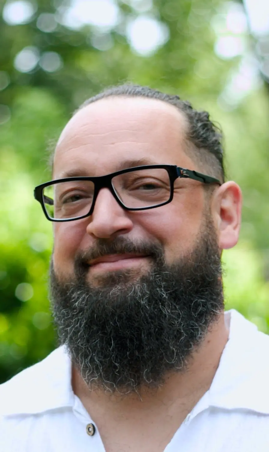 Portrait de Laurent Soler, souriant, en chemise blanche devant un fond de verdure